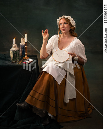Portrait of beautiful woman in vintage clothes sitting at table in dark room and sewing with dreamy smiling face again vintage green background Portrait of beautiful woman in vintage clothes sitting at table in dark room and sewing with dreamy smiling face again vintage green background 116396125