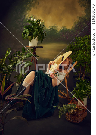 Little look. Elegant young woman in green velvet skirt and blouse, sitting on chair and hiding face behind big straw hat, again vintage actors with plants Little look. Elegant young woman in green velvet skirt and blouse, sitting on chair and hiding face behind big straw hat, again vintage actors with plants 116396179