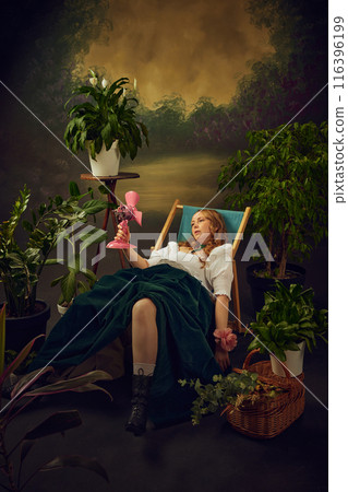 Super hot days. Woman in 18s century attire sitting tired in chair in green garden with small pink electric fan blowing on her, against vintage green background 116396199