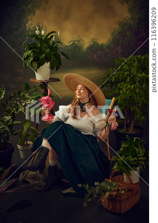 Smiling woman in period attire sitting on chair in garden with small pink electric fan blowing on face, drinking cocktail against vintage background. 116396209