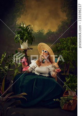 Portrait of elegant woman in period attire sitting on chair in green garden with small pink electric fan blowing on face, against vintage background. Hot summer day picnic gathering 116396217