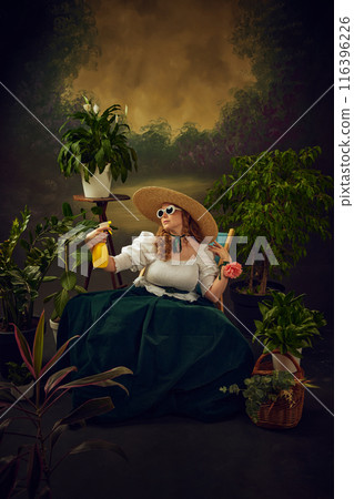 Summer hot day refreshment. Elegant young woman in velvet dress, white blouse, retro sunglasses and straw hat, sitting on chair in lush green garden and spraying water on herself 116396226