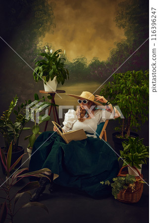 Portrait of elegant young woman in retro style glasses, straw hat, and velvet skirt, sitting on chair around lush green plants, and reading book against vintage background Portrait of elegant young woman in retro style glasses, straw hat, and velvet skirt, sitting on chair around lush green plants, and reading book against vintage background 116396247
