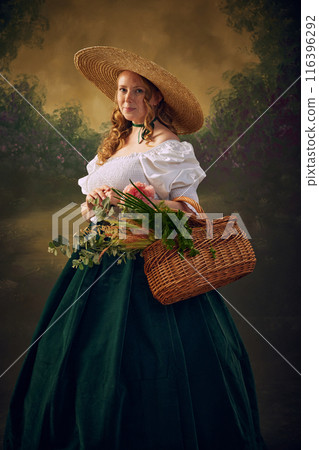 Portrait of woman in historical clothes, in large straw hat, white blouse, and green velvet skirt, holds wicker basket filled with greenery and flowers, set against vintage background 116396292