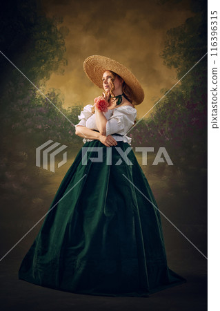 Portrait of elegant, beautiful young woman, medieval person in period attire, long skirt, blouse and hat, standing with thoughtful dreamy face against vintage green background 116396315