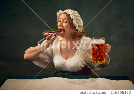 Young woman in medieval clothes taking giant bite of sandwich with fish and onion, holding beer mg, sitting at against vintage green background Young woman in medieval clothes taking giant bite of sandwich with fish and onion, holding beer mg, sitting at against vintage green background 116396330