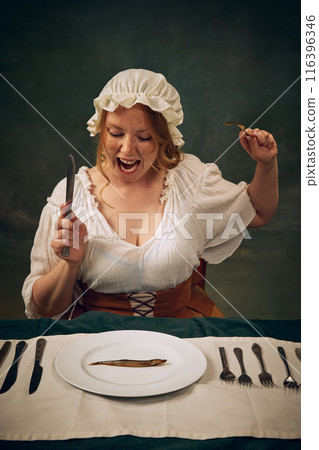 Emotional woman in medieval clothes, bonnet and white blouse sitting at table with knife and fork, emotionally looking on plate with little fish, set against vintage green background Emotional woman in medieval clothes, bonnet and white blouse sitting at table with knife and fork, emotionally looking on plate with little fish, set against vintage green background 116396346