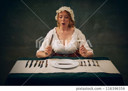 Emotional woman in historical attire, with bonnet and white blouse sitting at table with multiple knives and forks, looking on plate with single small fish. Vintage Green background 116396356