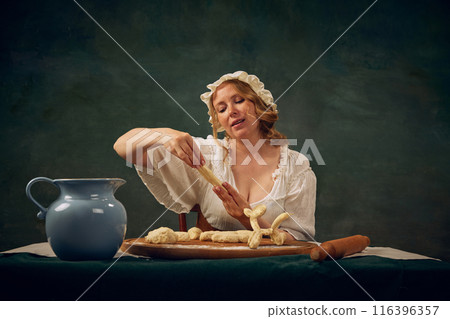 Portrait of beautiful, Medieval woman in period attire sitting at table and making pastry with freshly made dough, set against vintage green background 116396357