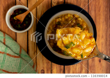 Close-up of a spoon with locro. Blurred background of a white bowl with typical Argentine cuisine Locro and the spicy sauce that accompanies this stew on a wooden table. Aerial view. 116398014