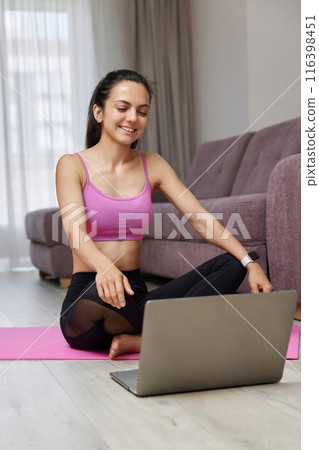 Happy Caucasian woman looking at laptop while sitting on pink fitness mat, ready for online training and workout at home. Online training. 116398451