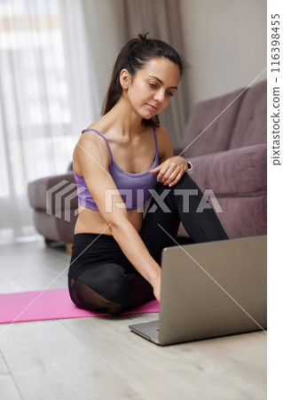 Happy sporty Caucasian woman looking at laptop while sitting on pink fitness mat, ready for online training and workout at home. 116398455