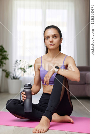 caucasian woman drinking water sitting on pink exercise mat at home 116398473