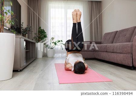 sporty woman working out lying on her back on pink mat lifting body and legs above head at home 116398494