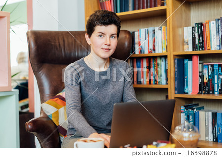 Confident middle aged female expert sits with laptop in leather armchair in cozy office with bookshelf background. 40s middle-age professional portrait of teacher, coach, mentor, therapist, counselor. 116398878