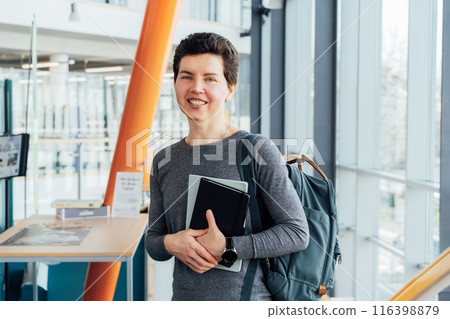 Smiling neutral gender middle-aged female person in casual clothes with backpack and books stands on staircase in modern building of university or education Centre. Adult students, Academic education. 116398879