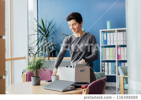 Satisfied newcomer middle aged neutral gender female company worker standing in office unpacking box with personal belongings at new workplace having first working day concept. New Carrier start. Satisfied newcomer middle aged neutral gender female company worker standing in office unpacking box with personal belongings at new workplace having first working day concept. New Carrier start. 116398888