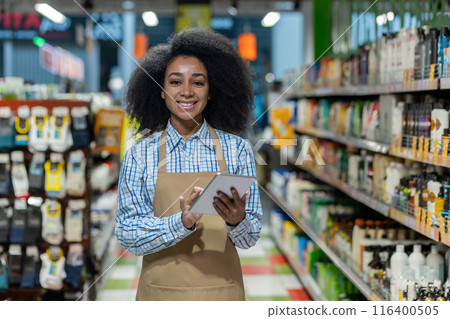 Female retail worker standing in store aisle holding tablet, smiling, wearing apron and checked shirt. Employee demonstrating a modern approach to inventory management and customer service 116400505