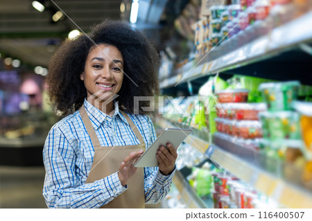 Confident supermarket employee using digital tablet while standing in front of dairy products aisle. Young woman working in retail, managing inventory ensuring product availability in grocery store Confident supermarket employee using digital tablet while standing in front of dairy products aisle. Young woman working in retail, managing inventory ensuring product availability in grocery store 116400507