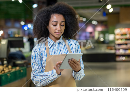 Retail worker using digital tablet in supermarket for inventory management. Employee wearing apron and checkered shirt standing in grocery store. Modern technology in retail industry. Retail worker using digital tablet in supermarket for inventory management. Employee wearing apron and checkered shirt standing in grocery store. Modern technology in retail industry. 116400509