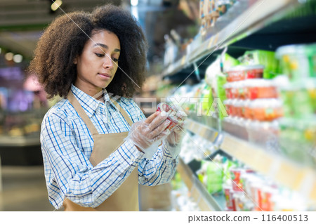 Supermarket employee checking product labeling on shelves at grocery store. Female retail worker wearing apron and gloves examining food packaging. Concept of retail, grocery shopping quality control Supermarket employee checking product labeling on shelves at grocery store. Female retail worker wearing apron and gloves examining food packaging. Concept of retail, grocery shopping quality control 116400513