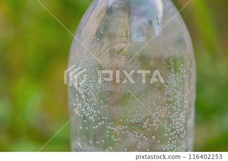 Air bubbles in the water that appeared inside a transparent plastic bottle in warm weather against the background of a green blurred background of vegetation. Air bubbles in the water that appeared inside a transparent plastic bottle in warm weather against the background of a green blurred background of vegetation. 116402253