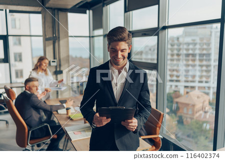 Smiling businessman in suit standing with clipboard at office on colleagues background Smiling businessman in suit standing with clipboard at office on colleagues background 116402374