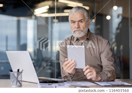 Senior businessman with beard using tablet seated at modern office desk with laptop, documents, and office supplies. Focused expression, professional attire, contemporary background 116403568