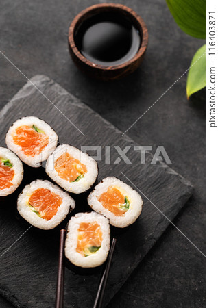 The concept of sushi maki with salmon, cucumber and nori on black slate boards with soy sauce.  116403871