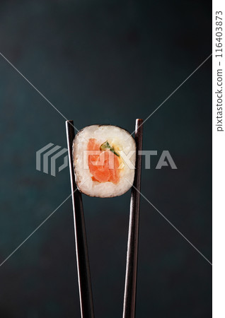 Chopsticks hold a sushi roll with salmon close-up on a dark background. Concept Japaness food 116403873