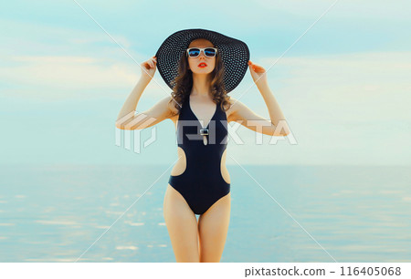 Summer beautiful young woman in bikini swimsuit, black hat, stylish girl posing on the beach on sea Summer beautiful young woman in bikini swimsuit, black hat, stylish girl posing on the beach on sea 116405068