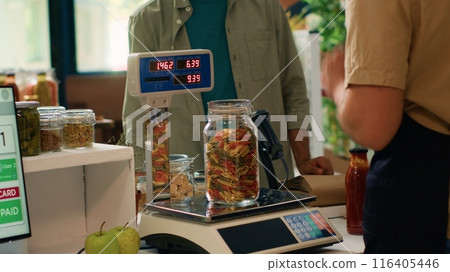 Salesperson in convenience store weighs fresh produce and groceries for middle eastern shopper by using digital scale at register. Retailer in local shop selling bulk pantry goods. Handheld shot. 116405446