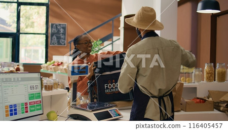 Vendor gives food order for delivery, putting homegrown organic veggies in backpack. African american woman courier coming to get bio supermarket merchandise for local customers. 116405457