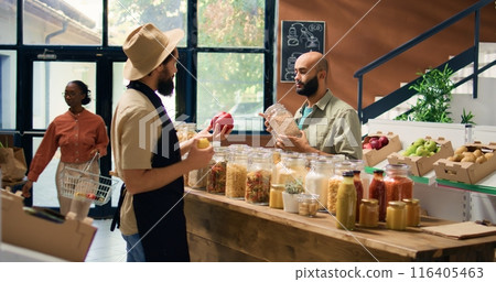 Vendor offers buyer bulk products, presenting middle eastern shopper selection of pasta, herbs and sauces. Young man buying organic produce at nearby eco friendly supermarket. Handheld shot. Vendor offers buyer bulk products, presenting middle eastern shopper selection of pasta, herbs and sauces. Young man buying organic produce at nearby eco friendly supermarket. Handheld shot. 116405463