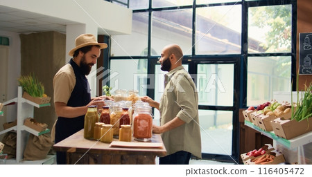 Shop owner shows pasta types to client, opening reusable containers filled with bulk items in natural bio grocery store. Vendor recommending organic additives free food goods. Shop owner shows pasta types to client, opening reusable containers filled with bulk items in natural bio grocery store. Vendor recommending organic additives free food goods. 116405472