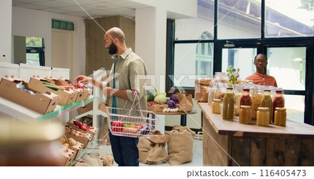 Buyer choosing ripe bio vegetables to buy for healthy sustainable dinner, looking at organic homegrown produce in crates at zero waste eco store. Man examining natural groceries. Tripod shot. Buyer choosing ripe bio vegetables to buy for healthy sustainable dinner, looking at organic homegrown produce in crates at zero waste eco store. Man examining natural groceries. Tripod shot. 116405473