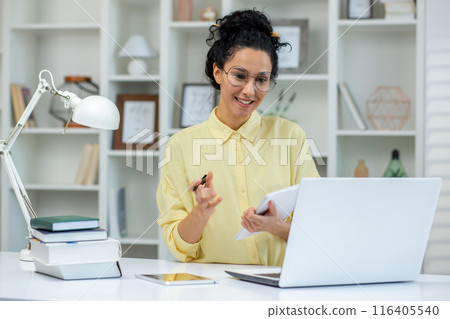 Smiling woman enjoying a study session at home. She is taking notes and using a laptop for online education. The cozy home office setup includes books, a lamp, and a tablet 116405540