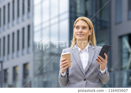 Confident businesswoman standing outside modern office building holding coffee cup and smartphone. Professional attire and focused expression depict success, business communication 116405607