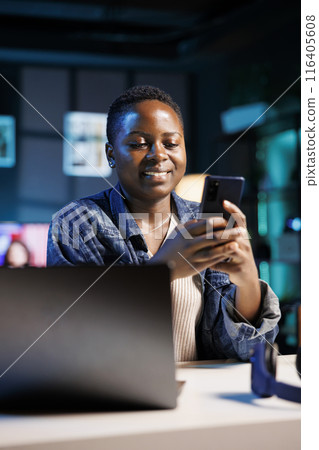 Black woman taking a break from working remotely on laptop and texting on cell phone using a social media app. African american freelancer at her home office desk chatting on mobile device. Black woman taking a break from working remotely on laptop and texting on cell phone using a social media app. African american freelancer at her home office desk chatting on mobile device. 116405608