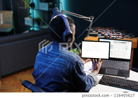 Black woman with headphones horizontally holding tablet with white screen. Female freelancer working from home, focused on digital device displaying blank chromakey mockup template. Black woman with headphones horizontally holding tablet with white screen. Female freelancer working from home, focused on digital device displaying blank chromakey mockup template. 116405627