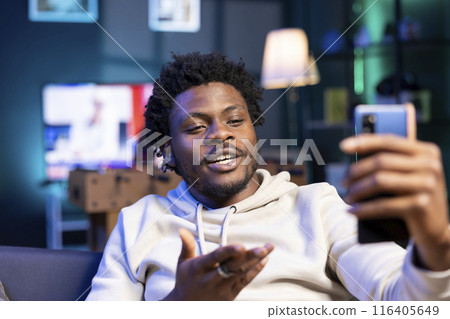 Smiling man speaking with close friend during teleconference conversation over the internet. Joyful person enjoying time together with mate in online videocall session while in cozy apartment 116405649