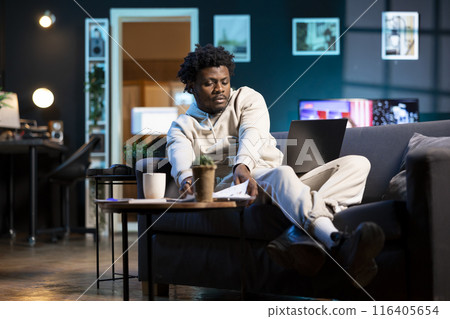 BIPOC man in apartment paying attention in elearning seminar teleconference with teacher, writing on notepad. Homeschooling student listening to lesson and checking presentation files 116405654