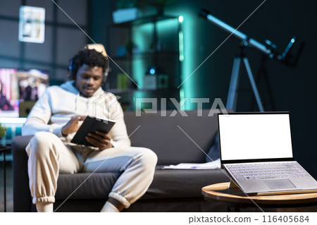 Man sitting on couch, reading eBook from tablet in apartment, focus on mockup laptop. Relaxed person wearing headphones, listening to audiobook next to isolated screen device on coffee table 116405684