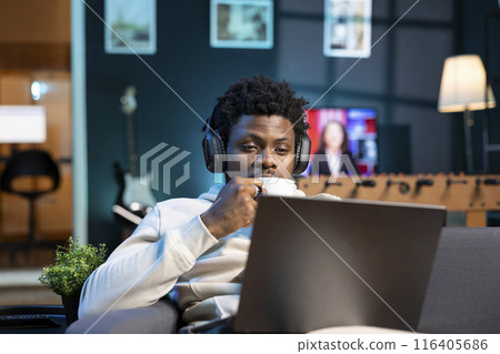 Self employed man drinking coffee and writing emails at home while listening music. African american remote employee doing b2b networking over internet, wearing headphones, enjoying hot beverage 116405686