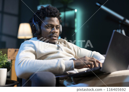 BIPOC man listening music while doing research for project using laptop, working from apartment office. Teleworker wearing headphones while inputting data on notebook, sitting down BIPOC man listening music while doing research for project using laptop, working from apartment office. Teleworker wearing headphones while inputting data on notebook, sitting down 116405692