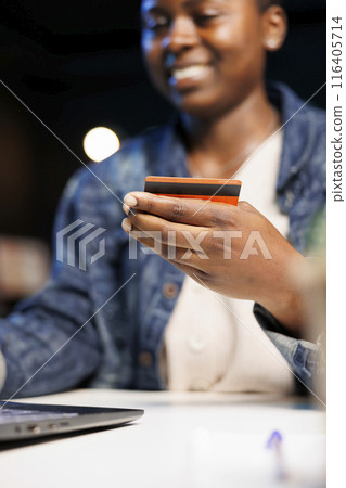 Smiling African American freelancer doing internet banking on her laptop, showcasing modern trend of remote work and financial management. Black lady grasps debit card for online purchases. Smiling African American freelancer doing internet banking on her laptop, showcasing modern trend of remote work and financial management. Black lady grasps debit card for online purchases. 116405714