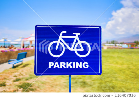 A vibrant blue traffic sign featuring a silhouette of a bicycle wheel parked against a sky backdrop with fluffy clouds. The font stands out against the natural landscape 116407036