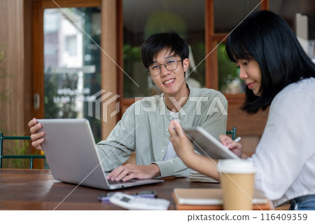 A positive Asian man is working remotely with his female colleague at a cafe, smiling at the camera. 116409359