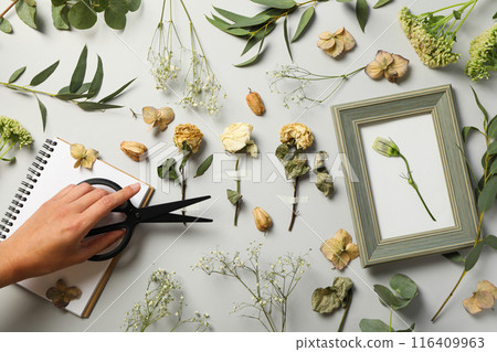 Dried flowers and plants, notepad, photo frame and hand with scissors on light background, top view 116409963