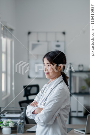 A confident female doctor in a white coat stands with arms crossed in a modern office, looking out the window. A confident female doctor in a white coat stands with arms crossed in a modern office, looking out the window. 116410987